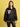 A woman with shoulder-length brown hair wears the Sanctuary Clothing x PEANUTS 75th Anniversary right on jacket black, featuring white trim and a "RIGHT ON!" Snoopy graphic on the left chest, posing before a yellow background.