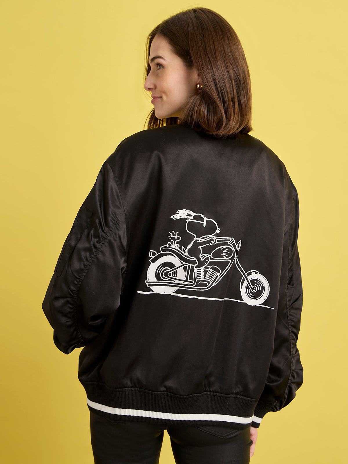 A woman with shoulder-length brown hair wears the right on jacket black by Sanctuary Clothing x PEANUTS 75th Anniversary, featuring a white line drawing of Snoopy riding a motorcycle on the back, standing against a yellow background.