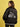 A woman with shoulder-length brown hair wears the right on jacket black by Sanctuary Clothing x PEANUTS 75th Anniversary, featuring a white line drawing of Snoopy riding a motorcycle on the back, standing against a yellow background.