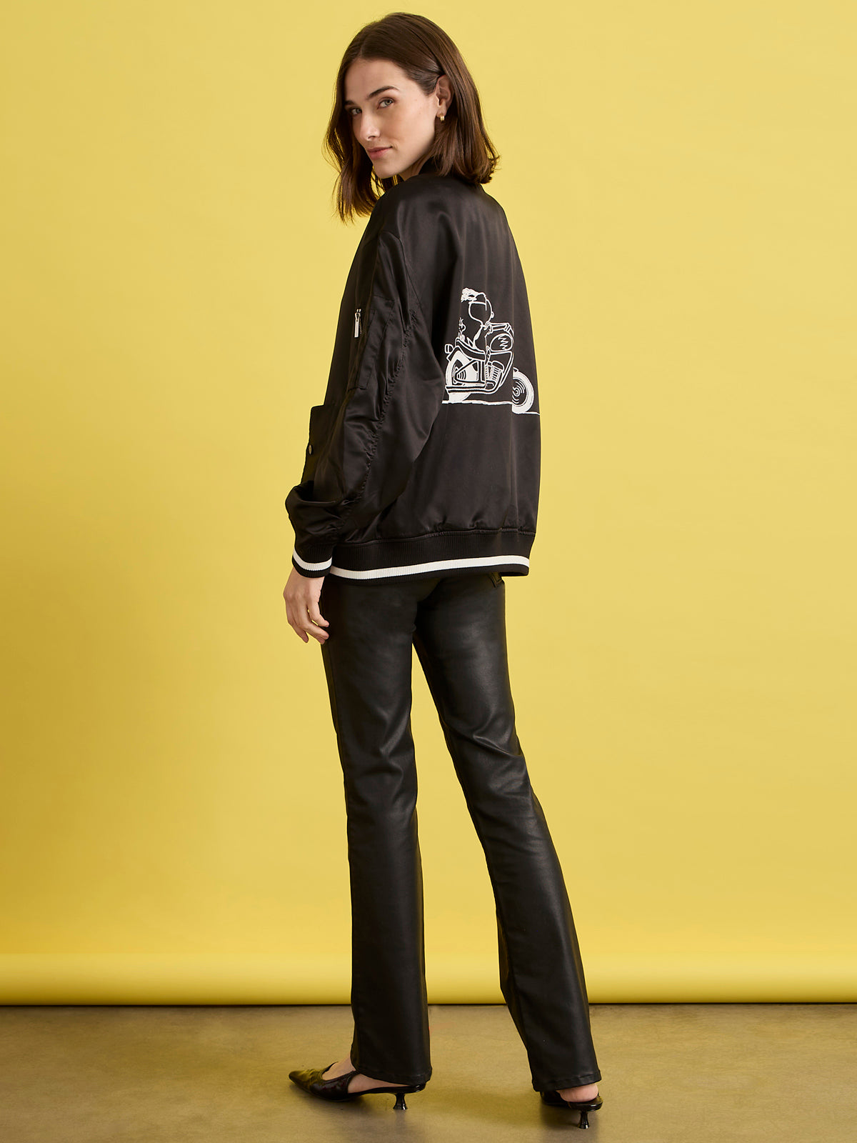 A woman stands in front of a yellow background, looking over her shoulder at the camera while wearing the Sanctuary Clothing x PEANUTS 75th Anniversary right on jacket black, paired with black leather pants and shoes.