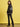 A woman stands in front of a yellow background, looking over her shoulder at the camera while wearing the Sanctuary Clothing x PEANUTS 75th Anniversary right on jacket black, paired with black leather pants and shoes.