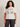 A woman with curly hair wears the "arent gonna keep quiet tee pink milk" from Sanctuary Clothing x PEANUTS 75th Anniversary, featuring comic characters and the quote, “We women aren't gonna keep quiet anymore, Chuck!” She smiles against a plain background.