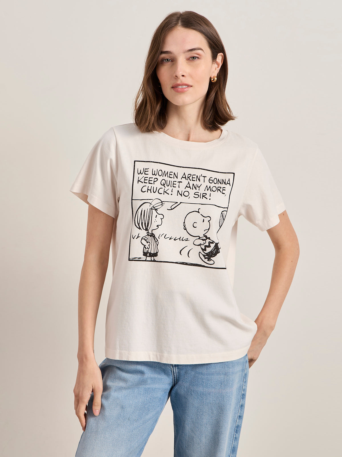 A woman with shoulder-length brown hair wears the Sanctuary Clothing x PEANUTS 75th Anniversary "arent gonna keep quiet tee pink milk," featuring a black-and-white Peanuts comic and bold text, with one hand in her pocket.