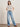 A woman poses casually in front of a light wall and brown floor, wearing the "arent gonna keep quiet tee pink milk" from Sanctuary Clothing x PEANUTS 75th Anniversary, paired with light blue wide-leg jeans and red shoes.