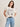 A woman with shoulder-length brown hair wears the pink milk "arent gonna keep quiet" tee from Sanctuary Clothing x PEANUTS 75th Anniversary, paired with blue jeans and a black belt. She stands smiling to the side, one hand in her pocket.