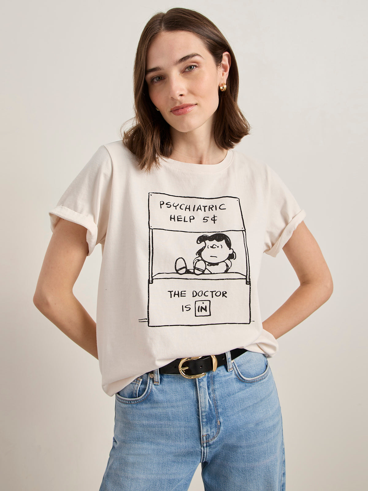 A woman with light skin and shoulder-length brown hair wears Sanctuary Clothing x PEANUTS 75th Anniversary's "the doctor is in tee collagen," a white T-shirt featuring the classic Peanuts "Psychiatric Help 5¢. The Doctor is IN." design.