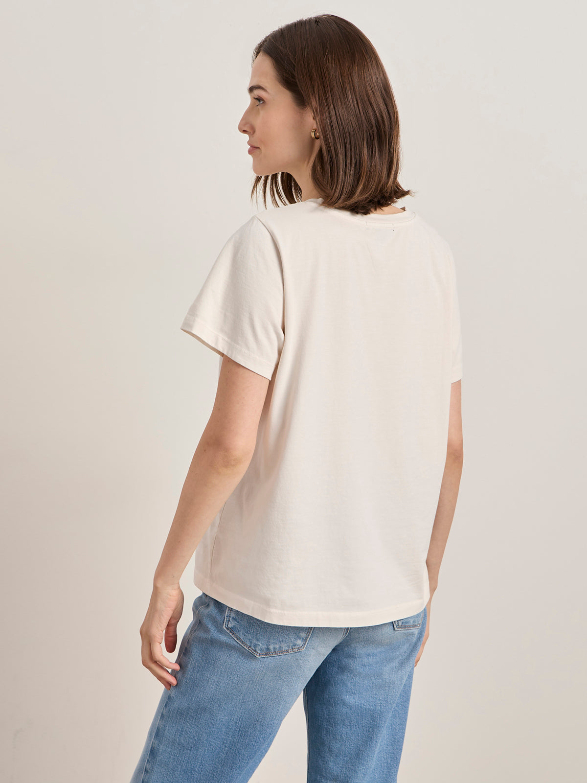 A woman with shoulder-length brown hair wears the Sanctuary Clothing x PEANUTS 75th Anniversary "the doctor is in" tee collagen (off-white, short-sleeved) and blue jeans, standing with her back to the camera against a plain light background.