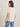 A woman with shoulder-length brown hair wears the Sanctuary Clothing x PEANUTS 75th Anniversary "the doctor is in" tee collagen (off-white, short-sleeved) and blue jeans, standing with her back to the camera against a plain light background.