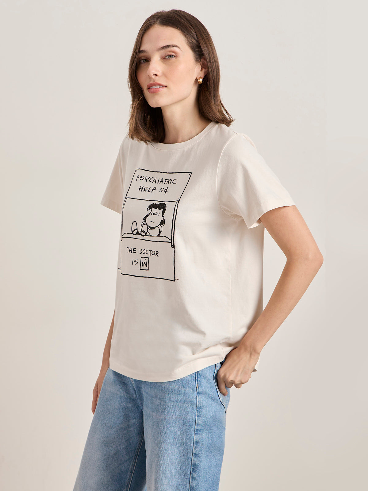 A woman wears the Sanctuary Clothing x PEANUTS 75th Anniversary "the doctor is in tee collagen," a cream T-shirt with Peanuts graphics and text, styled with light blue jeans against a plain background.