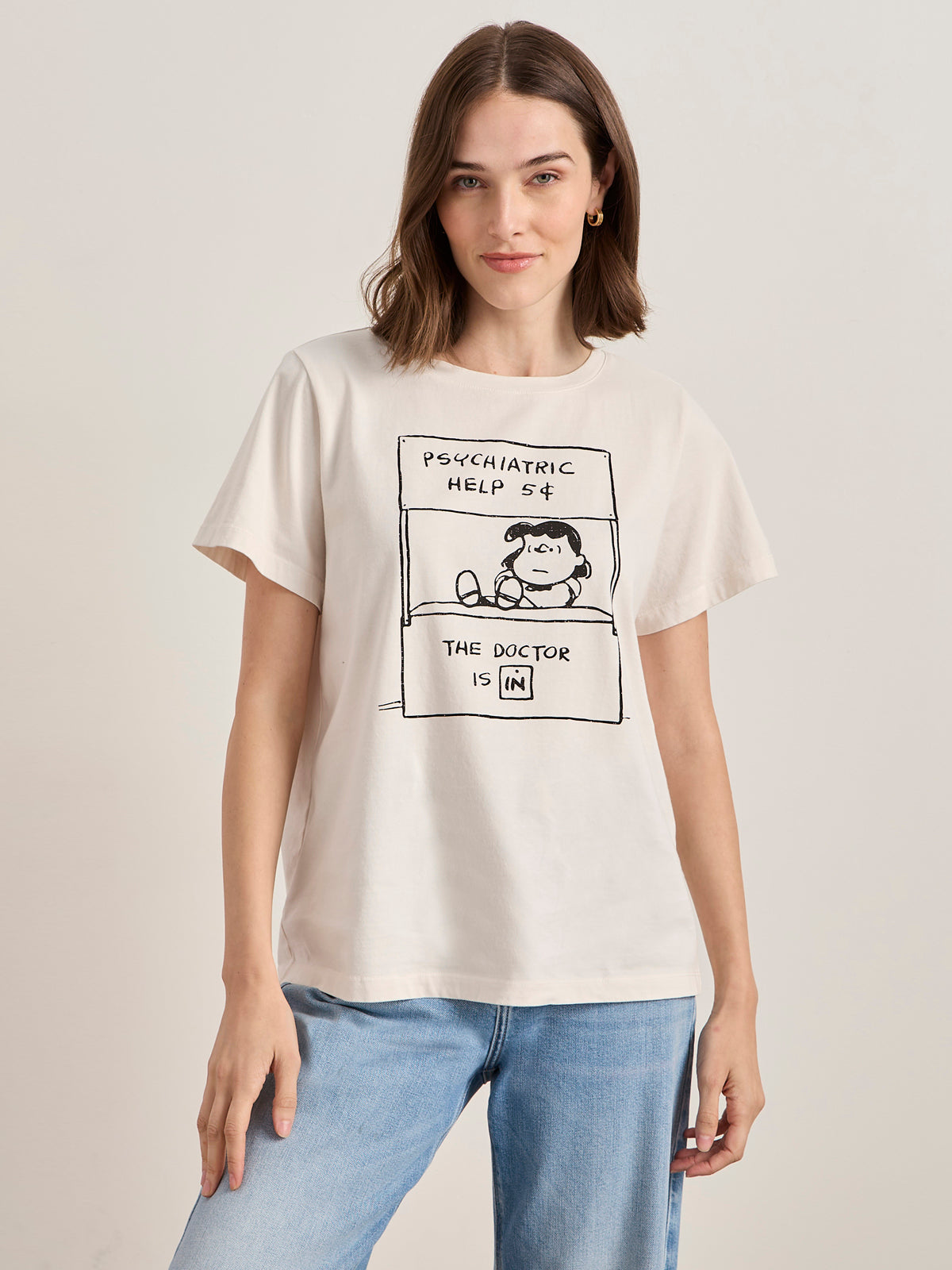 A woman with shoulder-length brown hair wears the Sanctuary Clothing x PEANUTS 75th Anniversary "the doctor is in tee collagen," a cream T-shirt featuring Lucy's "Psychiatric Help 5¢" and "The Doctor is In" text, paired with light blue jeans.