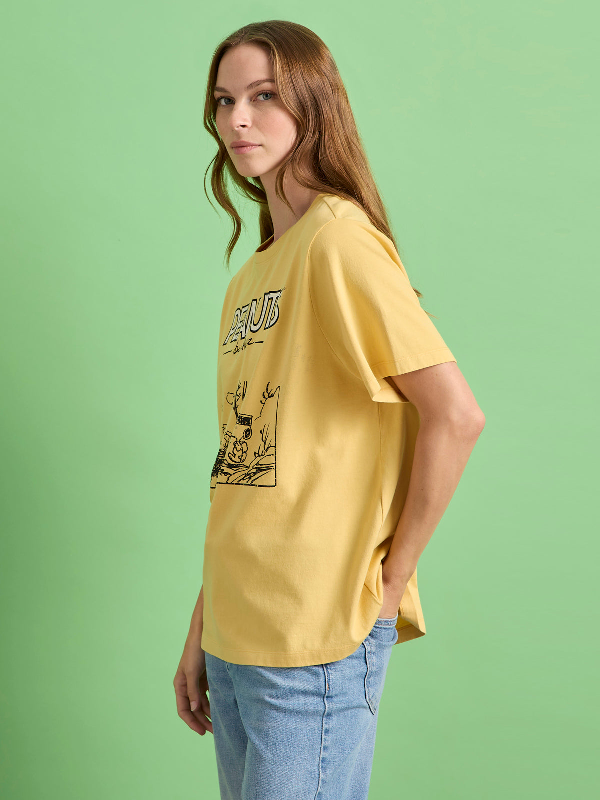 A woman with long brown hair wears the Sanctuary Clothing x PEANUTS 75th Anniversary marching tee in washed gold and light blue jeans, standing sideways against a plain light green background.