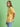 A woman with long brown hair wears the Sanctuary Clothing x PEANUTS 75th Anniversary marching tee in washed gold and light blue jeans, standing sideways against a plain light green background.
