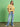 A woman stands against a green background wearing the Sanctuary Clothing x PEANUTS 75th Anniversary marching tee in washed gold, paired with light blue jeans, white sneakers, and a black belt. She has long hair and a relaxed expression.