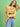A woman smiles against a green background, one hand in her hair, wearing Sanctuary Clothing x PEANUTS 75th Anniversary's marching tee in washed gold, paired with light blue jeans and a black belt.