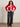A woman with curly hair smiles with hands on hips, wearing the Sanctuary Clothing x PEANUTS 75th Anniversary joe cool zip up sweater cherry lacquer, gray wide-leg jeans, a white top, and black ankle boots against a plain wall.