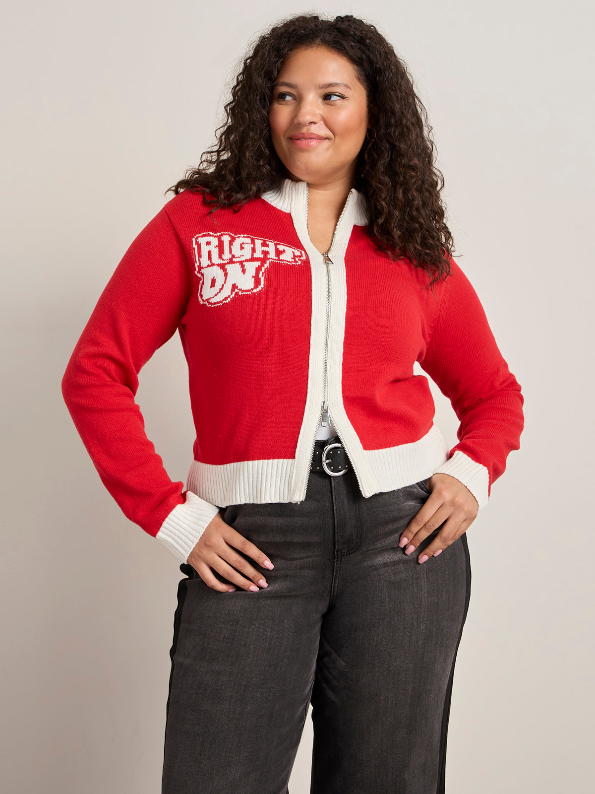 A woman with curly hair smiles, wearing the Sanctuary Clothing x PEANUTS 75th Anniversary joe cool zip up sweater in cherry lacquer, paired with high-waisted black jeans and a black belt. One hand is on her hip as she stands against a plain background.