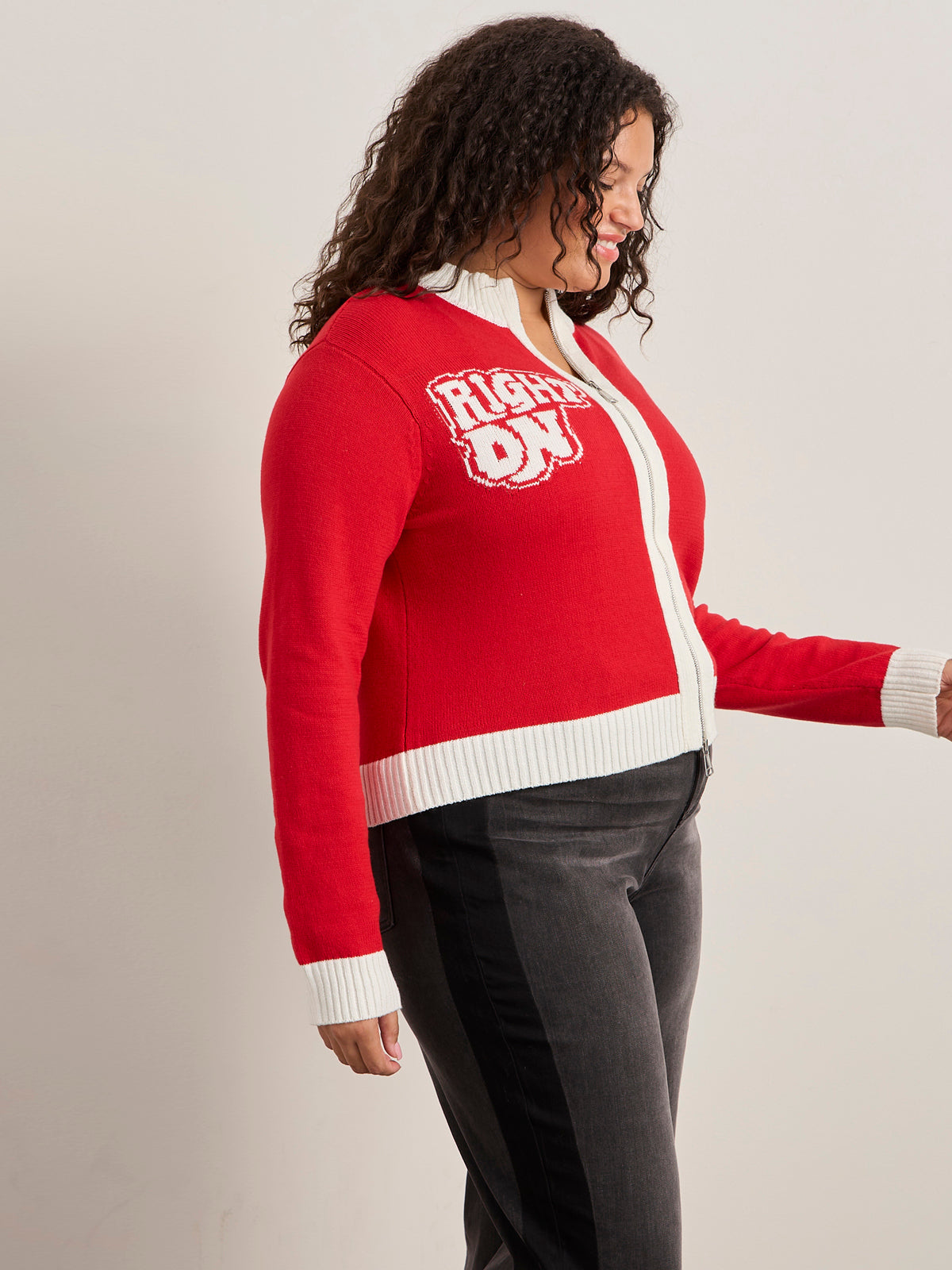A woman with curly hair wears the Sanctuary Clothing x PEANUTS 75th Anniversary joe cool zip up sweater in cherry lacquer, featuring white cuffs, collar, hem, and "PROUD OF" text. She pairs it with dark pants with a black side stripe, standing sideways and looking down.