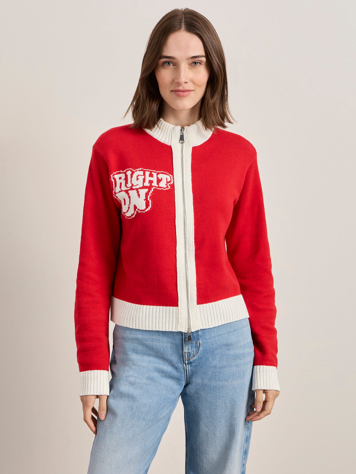 A woman with straight brown hair wears the Sanctuary Clothing x PEANUTS 75th Anniversary "joe cool zip up sweater cherry lacquer"—a red zip-up with white trim and "RIGHT ON" on the left chest—paired with light blue jeans against a plain light background.