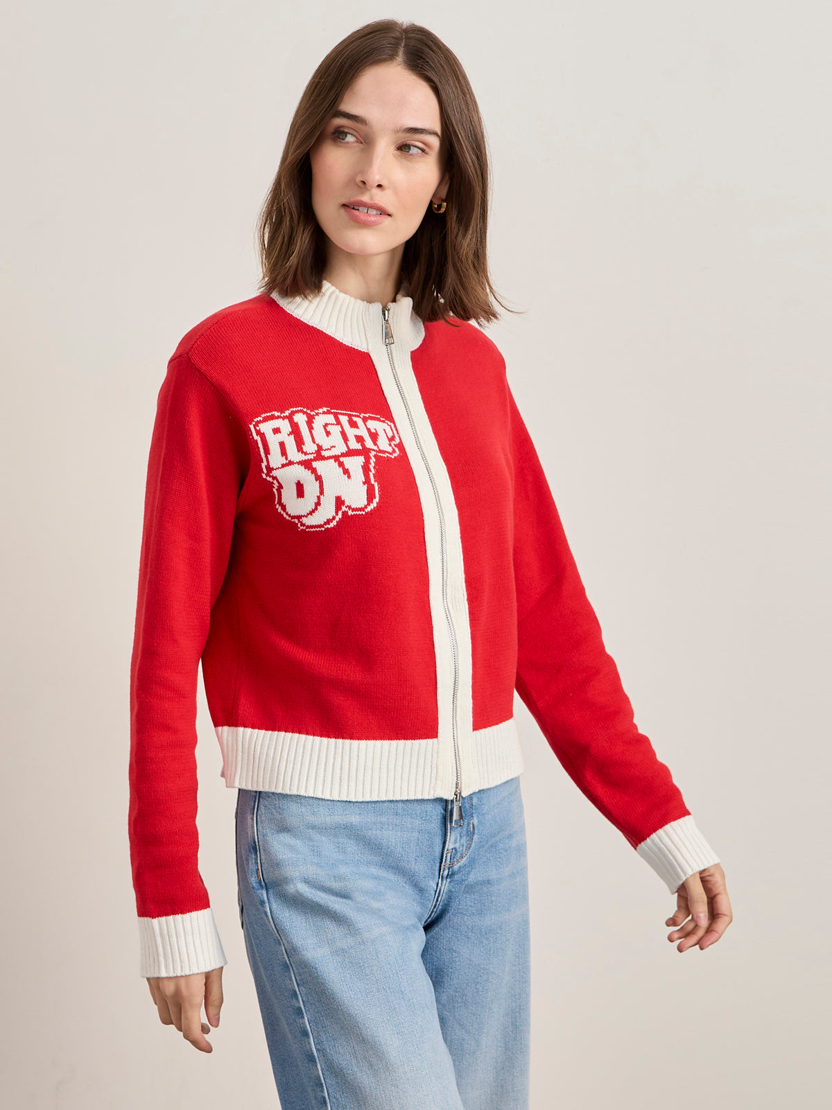 A woman in the Sanctuary Clothing x PEANUTS 75th Anniversary joe cool zip up sweater cherry lacquer, featuring white collar, cuffs, hem, and bold "RIGHT ON" text. She has shoulder-length brown hair and light blue jeans.