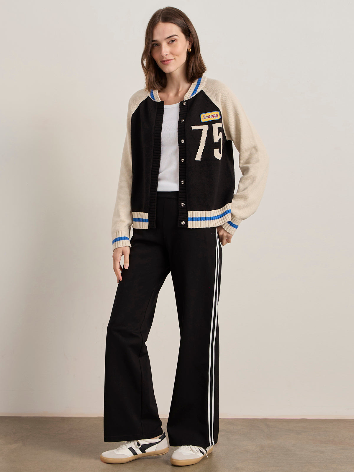A woman stands against a plain background wearing the Sanctuary Clothing x PEANUTS 75th Anniversary "75 bomber sweater black chalk," paired with a white top, black pants with white side stripes, and white sneakers. She smiles slightly with one hand in her pocket.