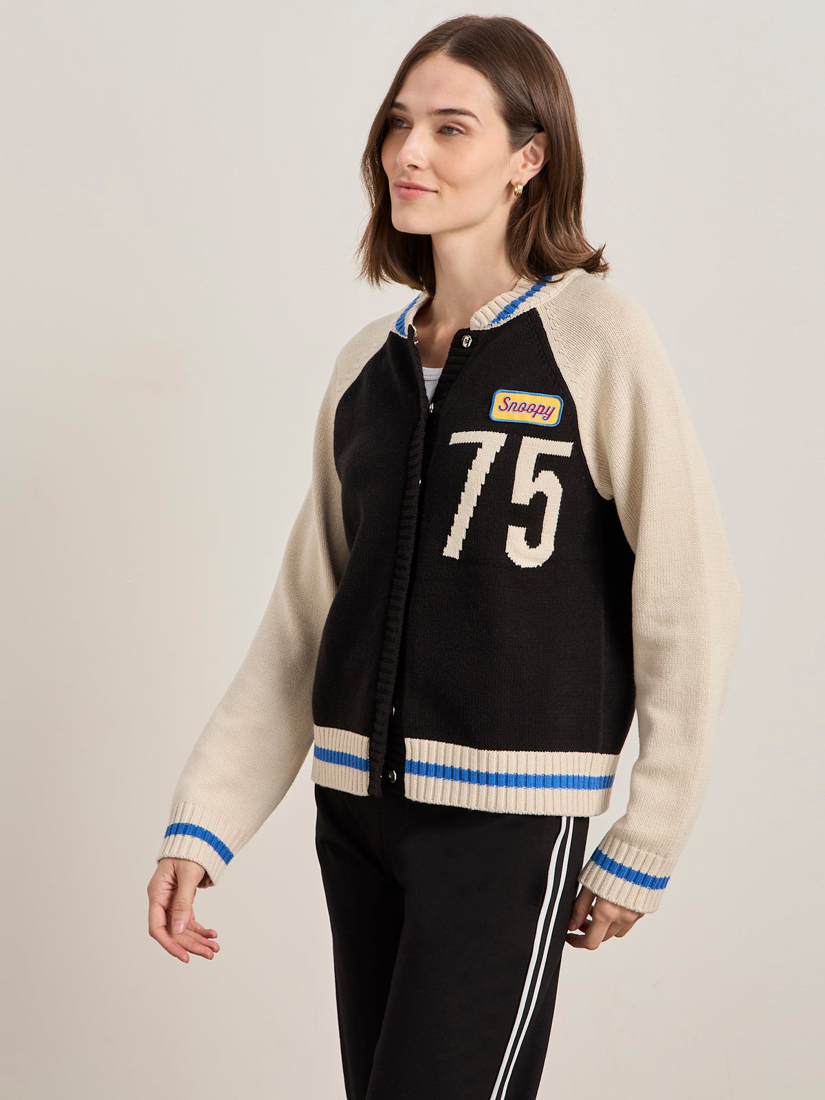A woman wears the Sanctuary Clothing x PEANUTS 75th Anniversary "75 bomber sweater black chalk," a varsity-style cardigan with "75" and "Snazzy" patches, blue and white striped cuffs and hem, standing against a plain background, looking to the side.