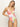 Against a light-colored background, a person with long hair poses in the "charming floral v wire mio white" swimsuit by Sanctuary Clothing, featuring large pink, green, and orange flowers.
