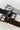 Three Sanctuary Clothing 'rowan belt chicory' belts in black, gray, and dark brown suede and leather, each with a large silver buckle, are arranged in a V-shape on a light background.