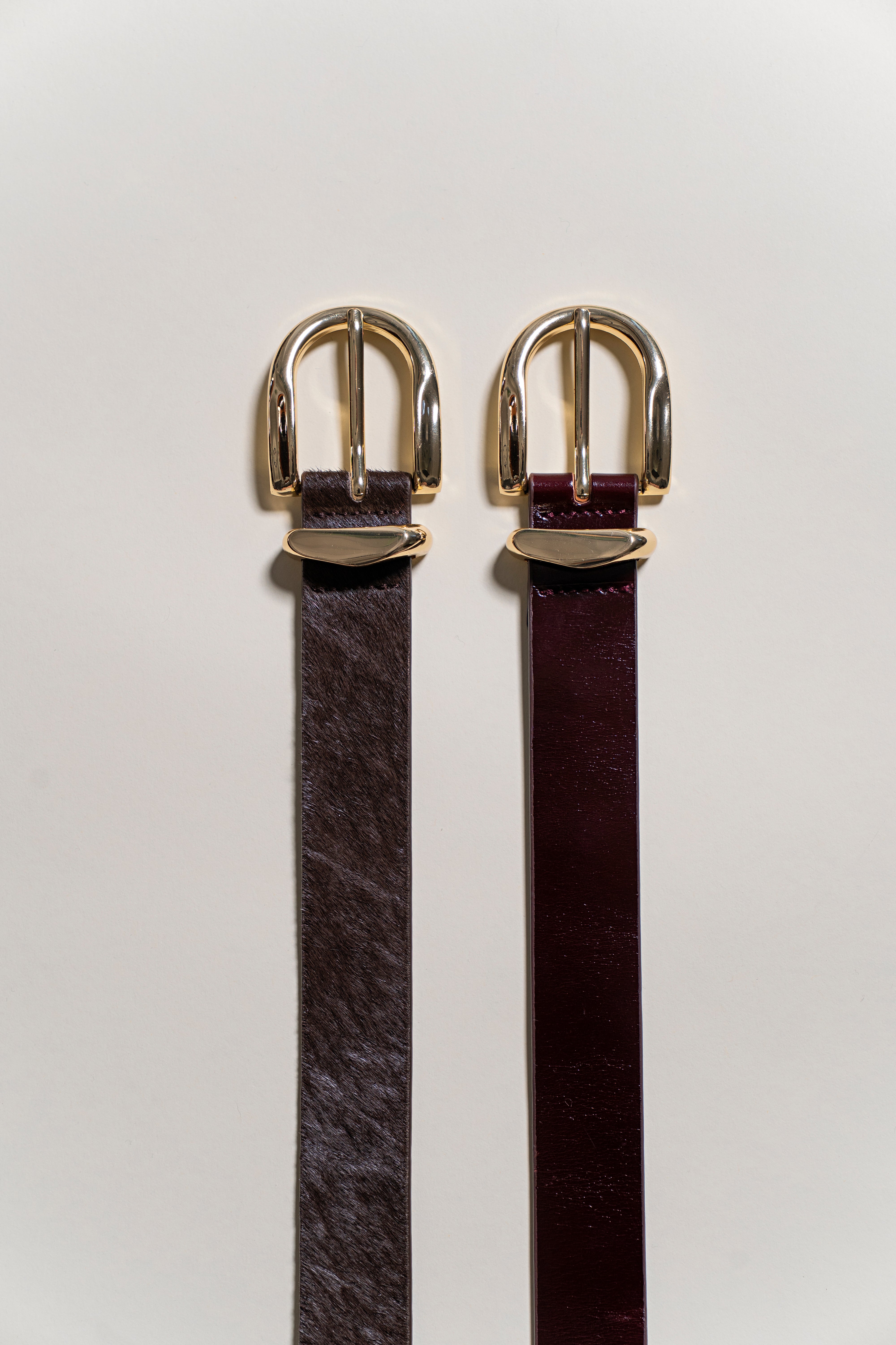 Two Sanctuary Clothing "all day belt dark crimson" belts rest side by side; one is dark brown with a textured finish, the other is burgundy with a smooth surface. Both have gold buckles and are displayed on a light background.