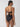 A woman with long dark hair stands against a plain, light background wearing Sanctuary Clothing's black refresh rib bikini bottom. The high-waisted design highlights her figure, and she faces away to showcase the tied back top.