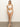 A person with long blonde hair stands barefoot on a plain indoor floor, wearing Sanctuary Clothing's "refresh rib bandeau white" top paired with high-waisted bottoms. The background is a simple, light-colored wall.
