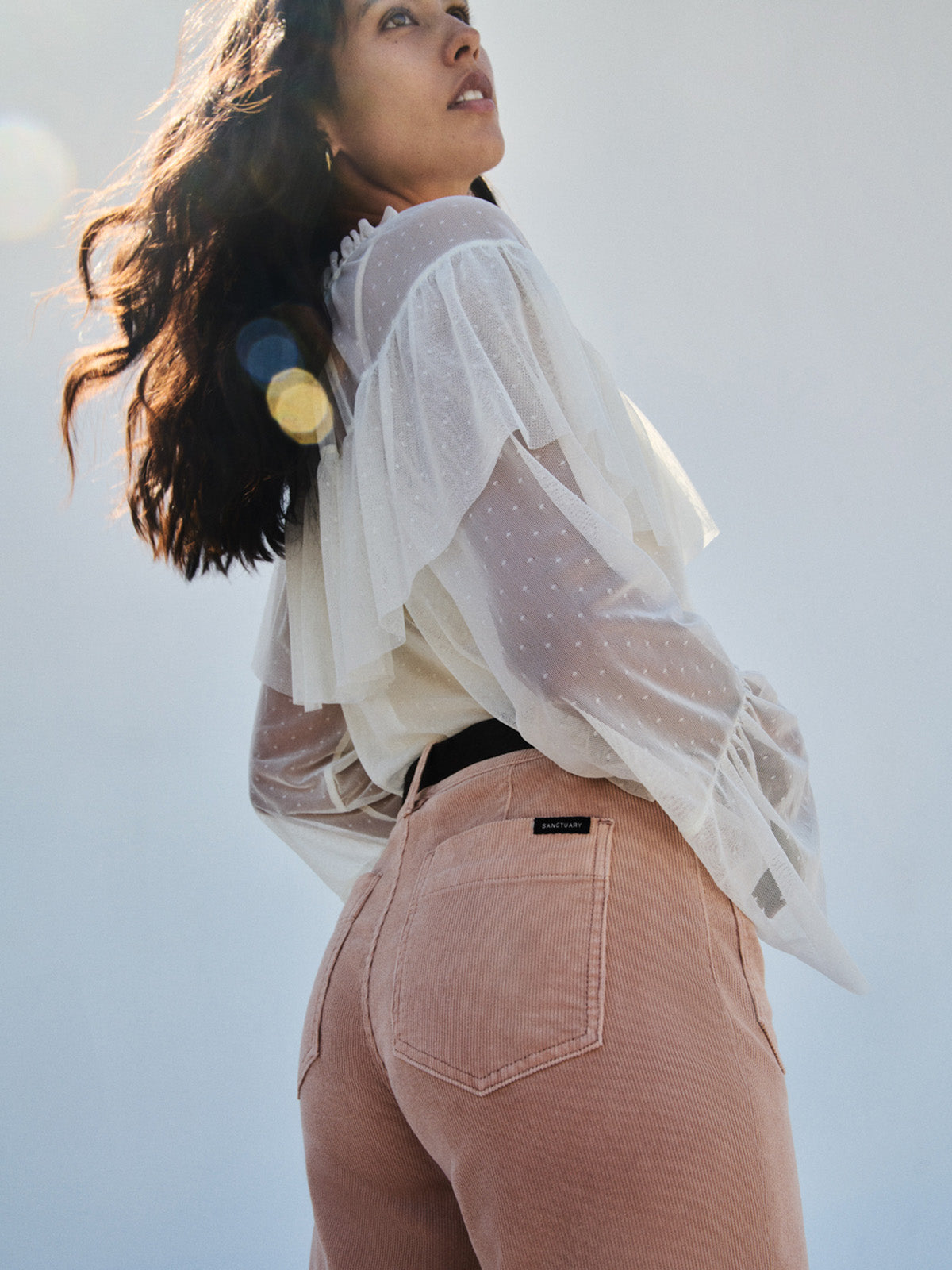 A woman with wavy hair stands outdoors in sunlight, wearing a sheer white blouse with ruffled sleeves and Sanctuary Clothing's corduroy marine pant in dusty rose.