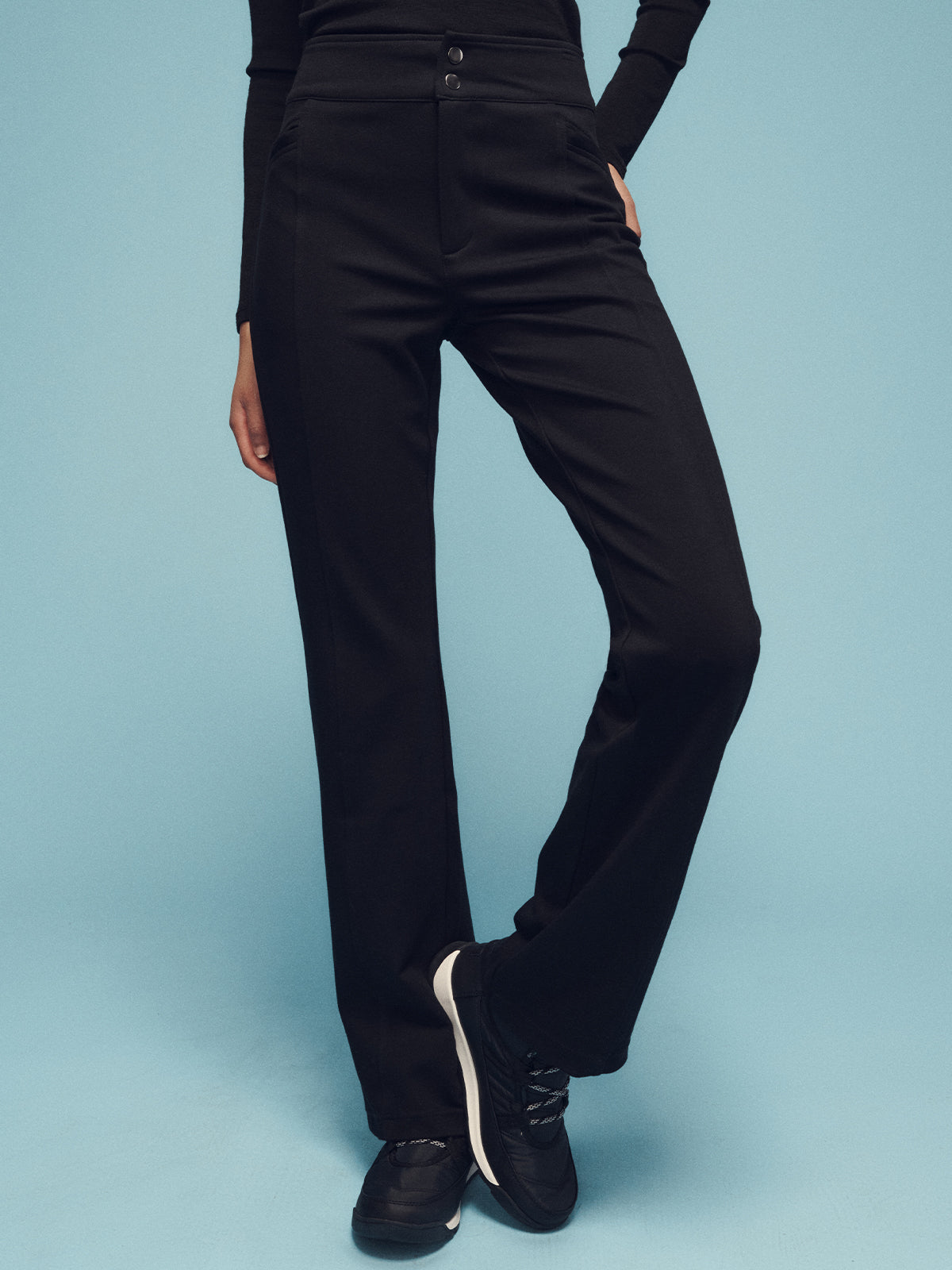 A person wearing the Ski Lodge Pants in black by Sanctuary Clothing stands against a light blue background. Their hands are in their pockets, and they are wearing a long-sleeved black top along with black sneakers.