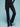 A person wearing the Ski Lodge Pants in black by Sanctuary Clothing stands against a light blue background. Their hands are in their pockets, and they are wearing a long-sleeved black top along with black sneakers.