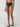 A person models the Sanctuary Clothing refresh rib bikini bottom black, paired with chic black sandals featuring beige straps against a plain background. The image highlights the striped detail on the bikini fabric and showcases the lower half of the body.