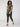 A person with curly dark hair is walking while wearing a camouflage T-shirt and the Runway Legging Black from Sanctuary Clothing's Inclusive Collection. They are also sporting transparent-heeled sandals and are looking to the side. The background is plain and light-colored.