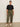A woman stands indoors against a plain light wall, wearing the Sanctuary Clothing shirred waist dolman tee in black (extended sizing). She pairs it with olive green cargo pants, a black belt, white sneakers with black stripes, and has her hair pulled back.
