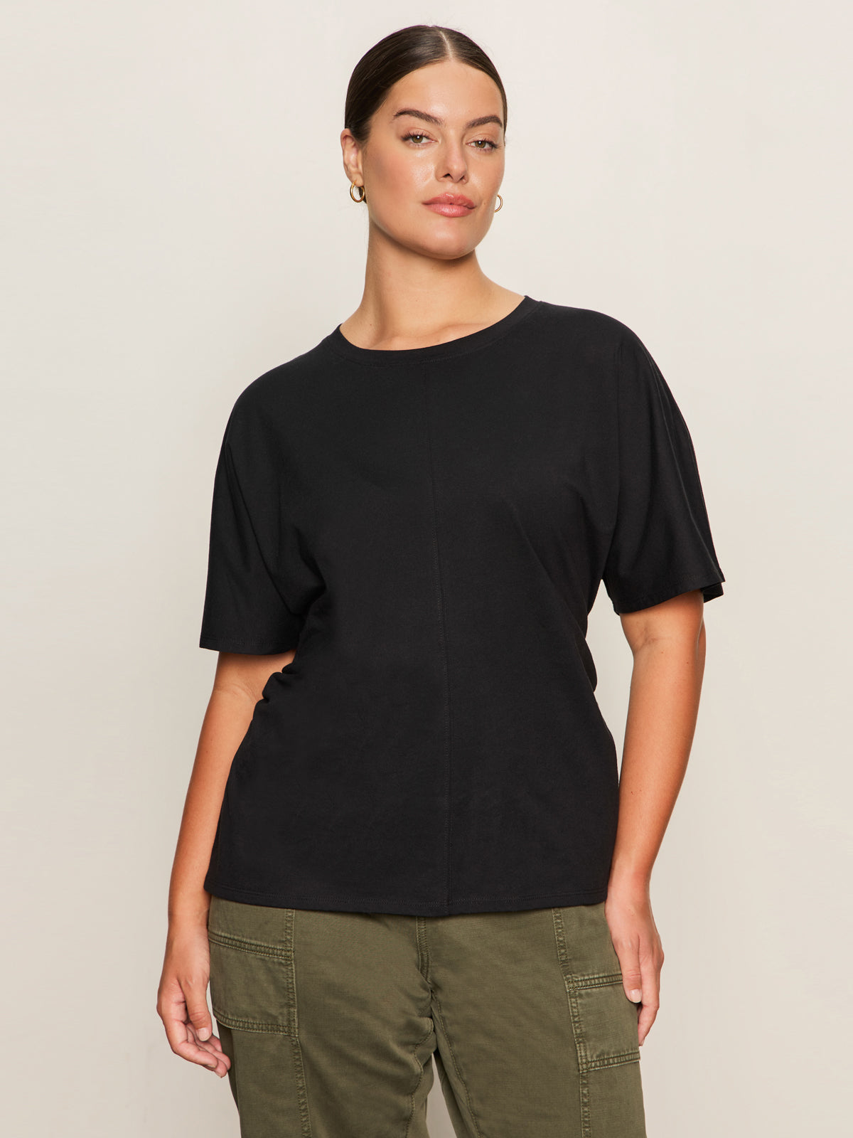 A woman with dark hair tied back wears the Sanctuary Clothing shirred waist dolman tee in black (extended sizing), paired with olive green cargo pants, standing against a plain light background and looking confidently at the camera.