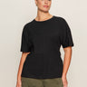 A woman with dark hair tied back wears the Sanctuary Clothing shirred waist dolman tee in black (extended sizing), paired with olive green cargo pants, standing against a plain light background and looking confidently at the camera.