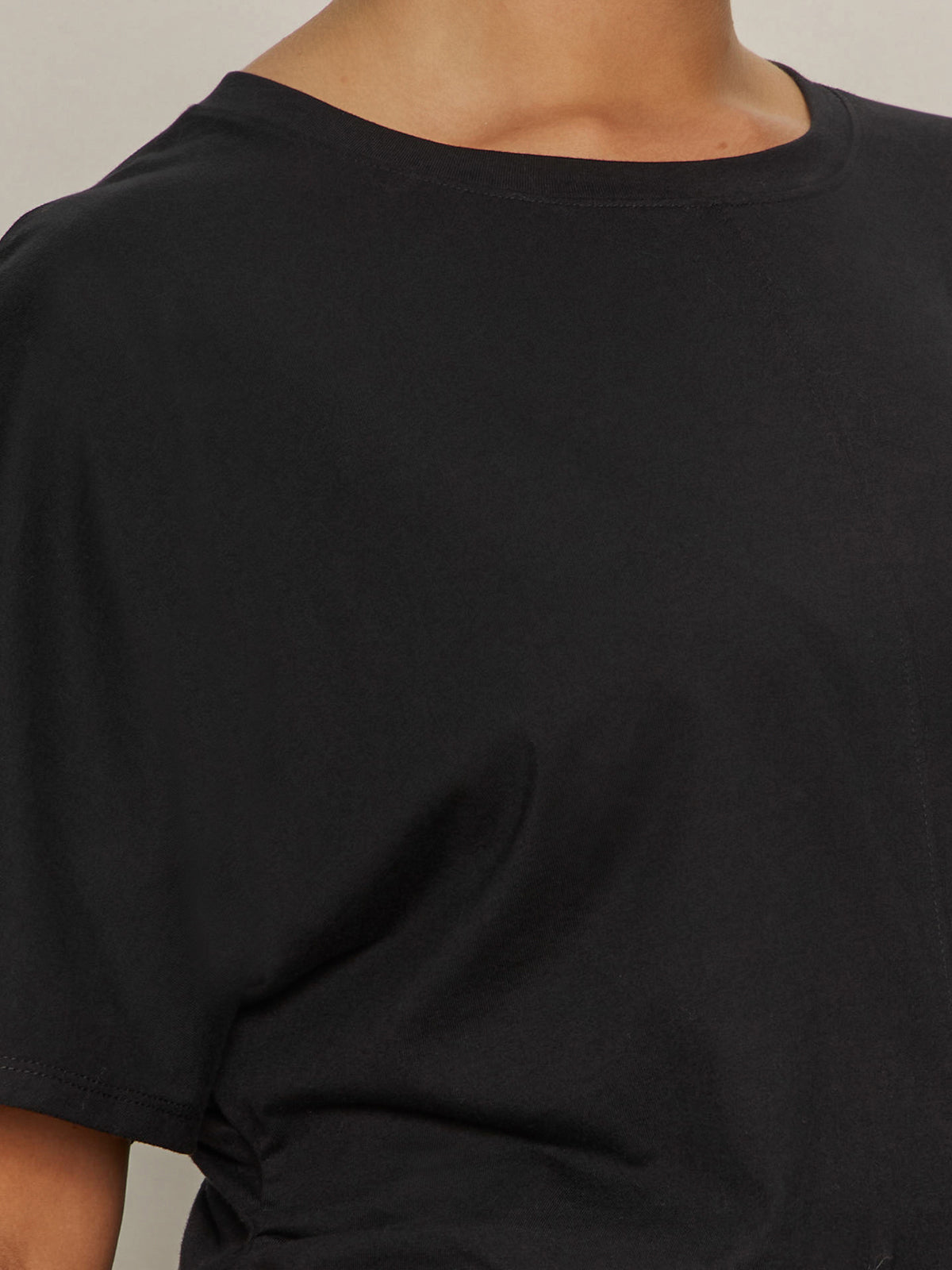A close-up of a person wearing the Sanctuary Clothing shirred waist dolman tee in black (extended sizing); only the upper chest, neck, and part of one arm are visible.