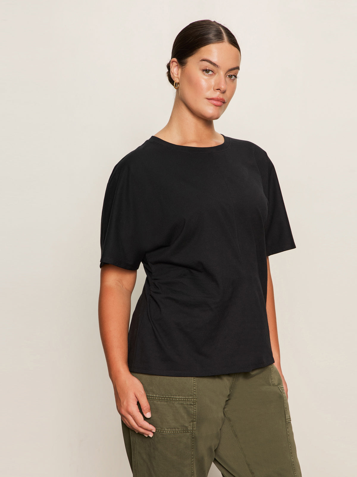 A woman with dark hair tied back wears Sanctuary Clothing's shirred waist dolman tee in black (extended sizing) and olive green cargo pants, standing against a plain light background and looking confidently at the camera.