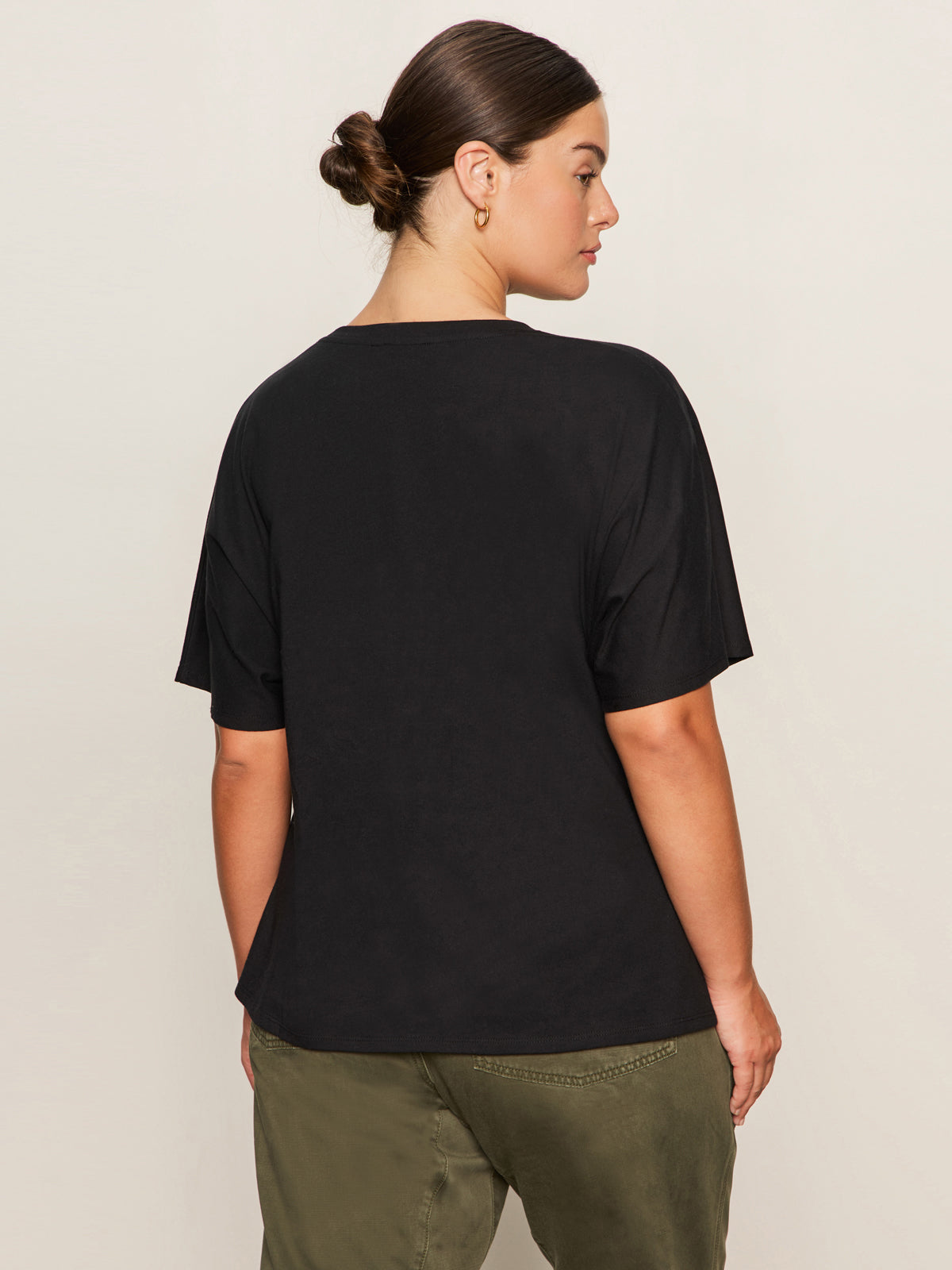 A woman with brown hair in a bun faces away from the camera, wearing the Sanctuary Clothing shirred waist dolman tee in black (extended sizing) and olive green pants against a plain light background.