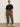 A woman poses confidently in Sanctuary Clothing's boyfriend utility pant kalamata (extended sizing), styled with a loose black tee, black-belted waist, and white sneakers. Her hair is pulled back as she stands against a plain wall.