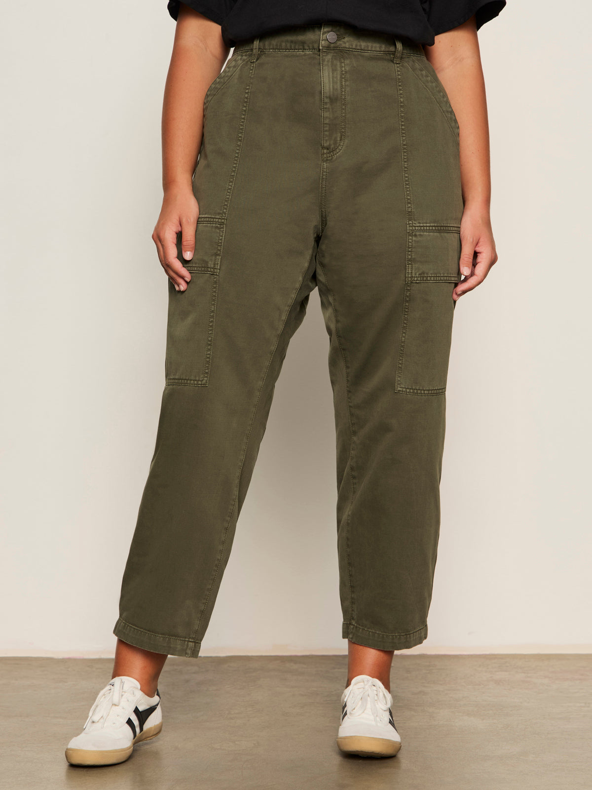 A person stands against a plain light background wearing Sanctuary Clothing's boyfriend utility pant kalamata extended sizing with large front pockets, paired with a black t-shirt and white sneakers featuring black accents. Their arms are relaxed by their sides.