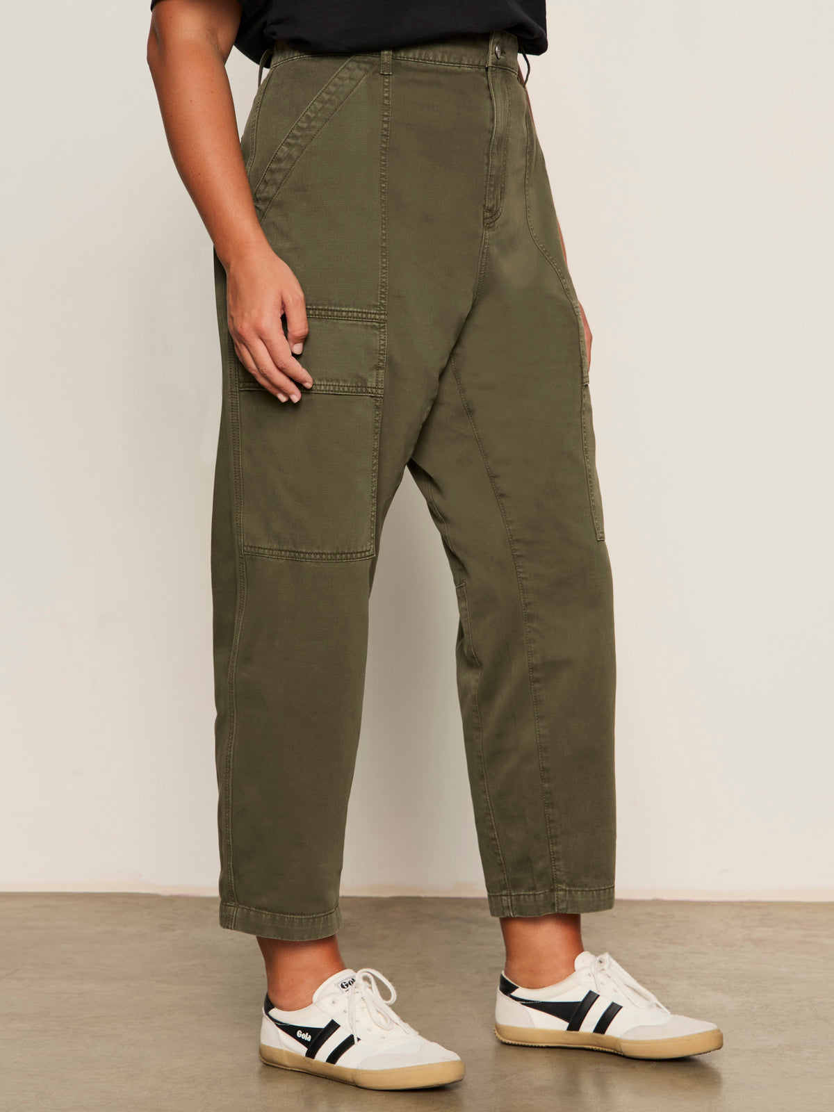 A person stands indoors, partially visible from shoulder to ankle, wearing Sanctuary Clothing's boyfriend utility pant in kalamata (extended sizing) and white sneakers with black stripes. Their left hand rests by their side.
