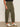 A person stands indoors, partially visible from shoulder to ankle, wearing Sanctuary Clothing's boyfriend utility pant in kalamata (extended sizing) and white sneakers with black stripes. Their left hand rests by their side.