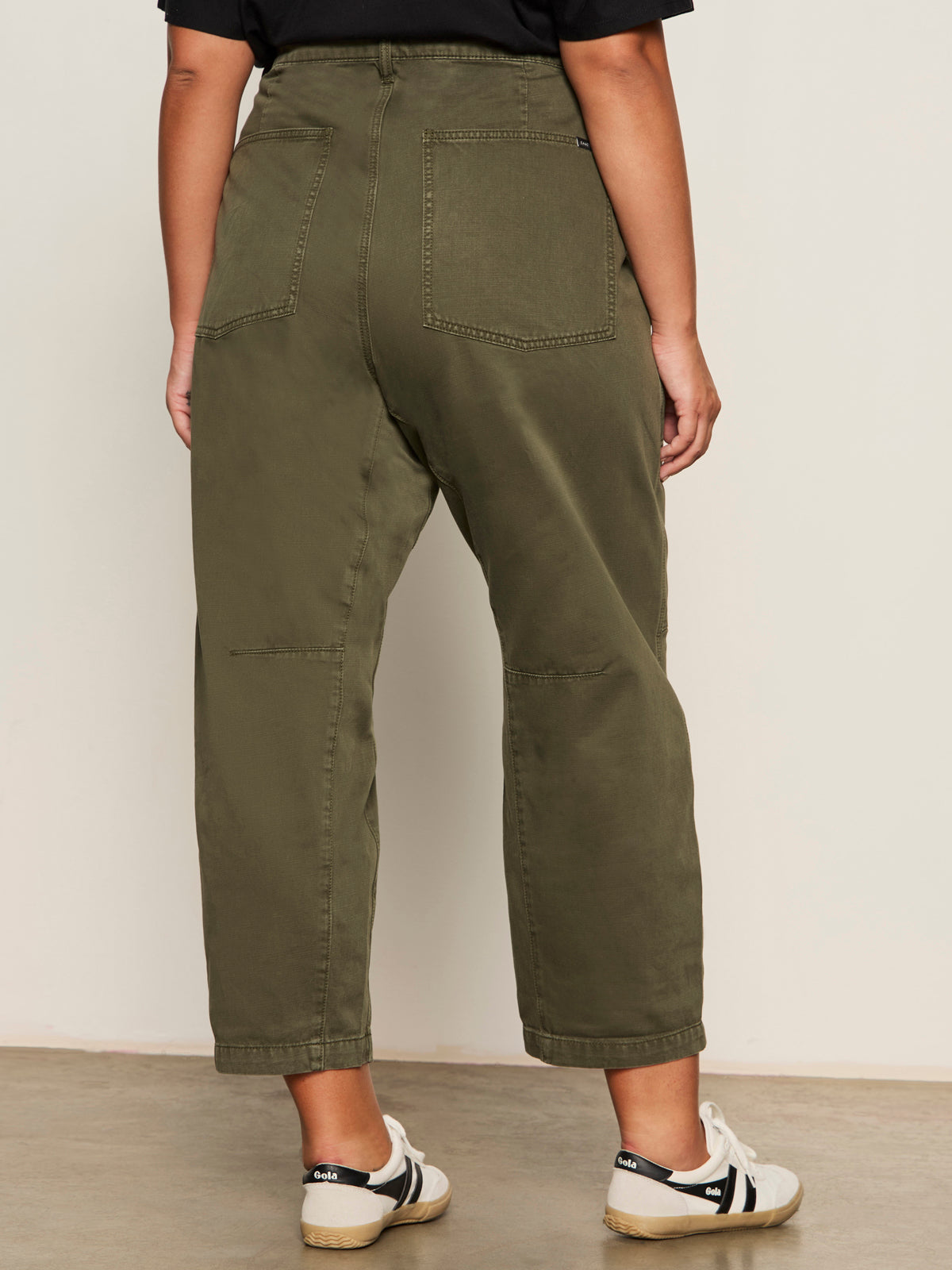 Indoors, a person faces away wearing Sanctuary Clothing's boyfriend utility pant kalamata (extended sizing) with large back pockets, paired with a black t-shirt and white sneakers featuring black stripes.