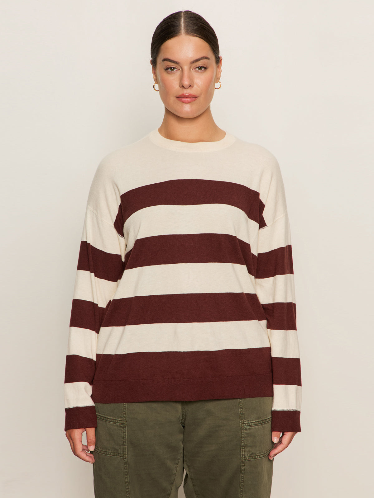 A person with dark hair tied back is wearing the Sanctuary Clothing 24/7 Popover Chicory Light Oat sweater, featuring cream and maroon stripes, paired with olive green pants against a plain light background.