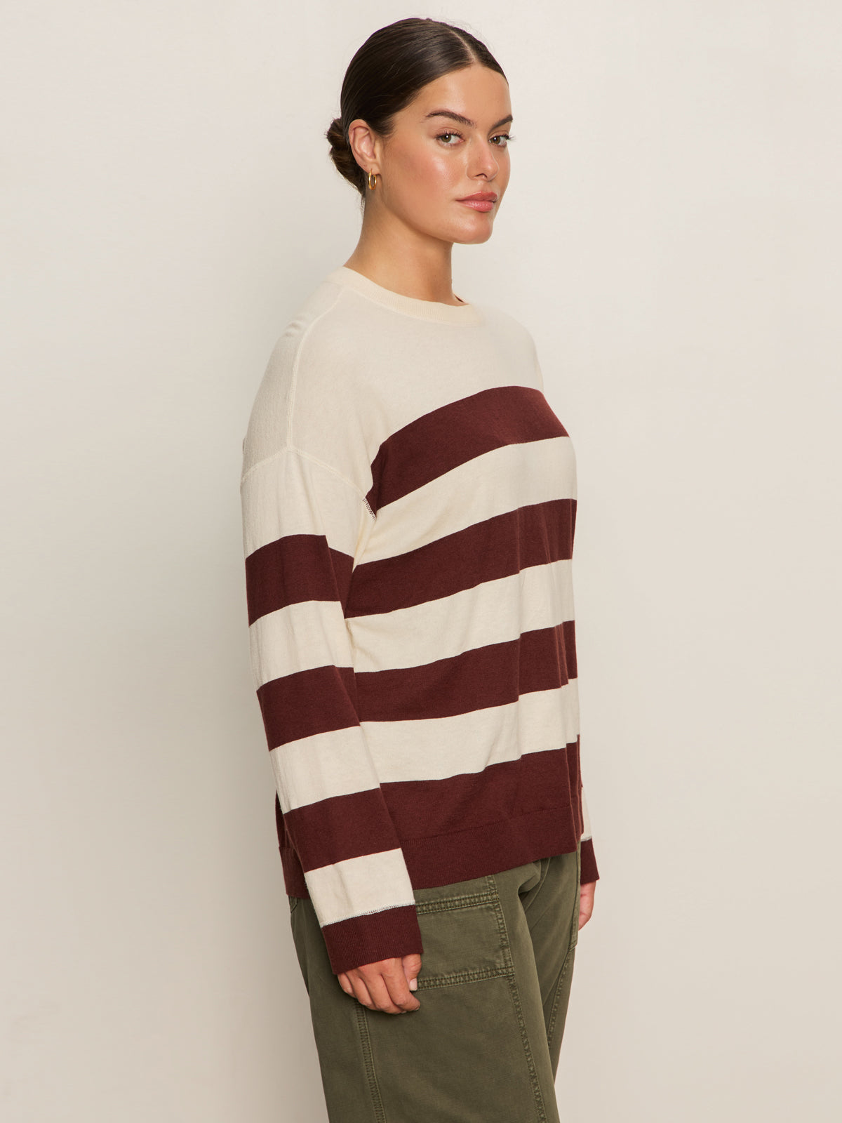 A person stands sideways against a light background, wearing the Sanctuary Clothing 24 7 Popover Chicory Light Oat sweater in extended sizing with olive green pants. Their dark hair is pulled back and they have a neutral expression.