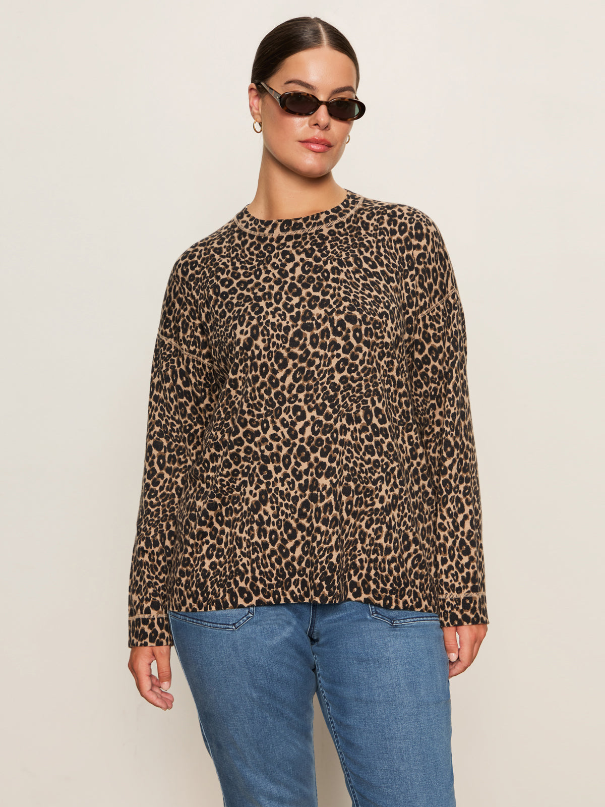 A woman stands confidently against a plain light background, wearing Sanctuary Clothing's 24 7 Popover French Kitty top and black sunglasses with blue jeans.