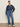 A woman with her hair tied back confidently poses in Sanctuary Clothing's personal best blouse moonshadow extended sizing, paired with high-waisted blue jeans and white sneakers with black accents against a plain background.